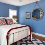 Bedroom design includes a bold blue and white rug to anchor this vintage bed frame painted in a cherry red metallic finish. Wall painted in Sherwin Williams sporty blue creates a dramatic backdrop. Custom pillows in a mix of fabrics combine the red and blue and create a curated look. Coastal artwork and table lamps complete the decor.
