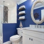 Coastal pool bath with navy walls and white shaker cabinets. A white rope mirror and nautical vanity lights add to the coastal decor.